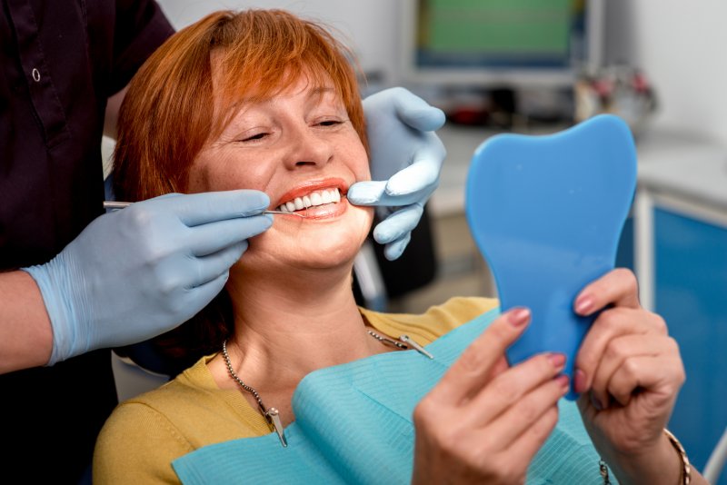 Woman with dental implants smiling and looking into hand-held mirror in dentist's chair.
